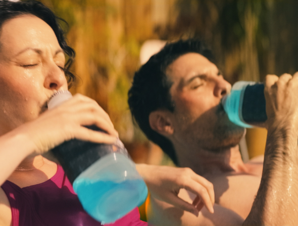 A mom and dad sit in a kiddie pool drinking Pedialyte® to hydrate during the summer heat.