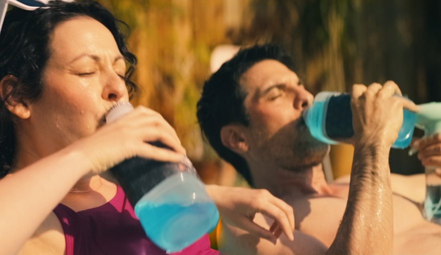 A mom and dad sit in a kiddie pool drinking Pedialyte® to hydrate during the summer heat.
